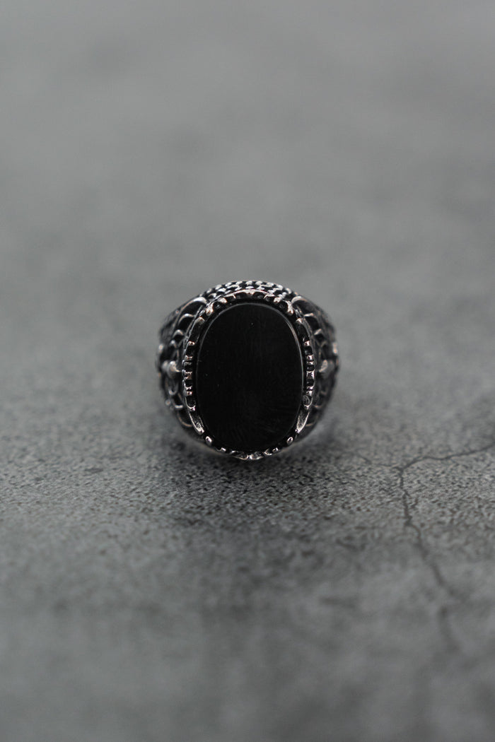 A close-up of the Royal Darkness Ring by Relentless Betrayal, a stainless steel ring with an oval black stone, intricate detailing, and a striking black finish on the band, displayed on a textured gray surface.