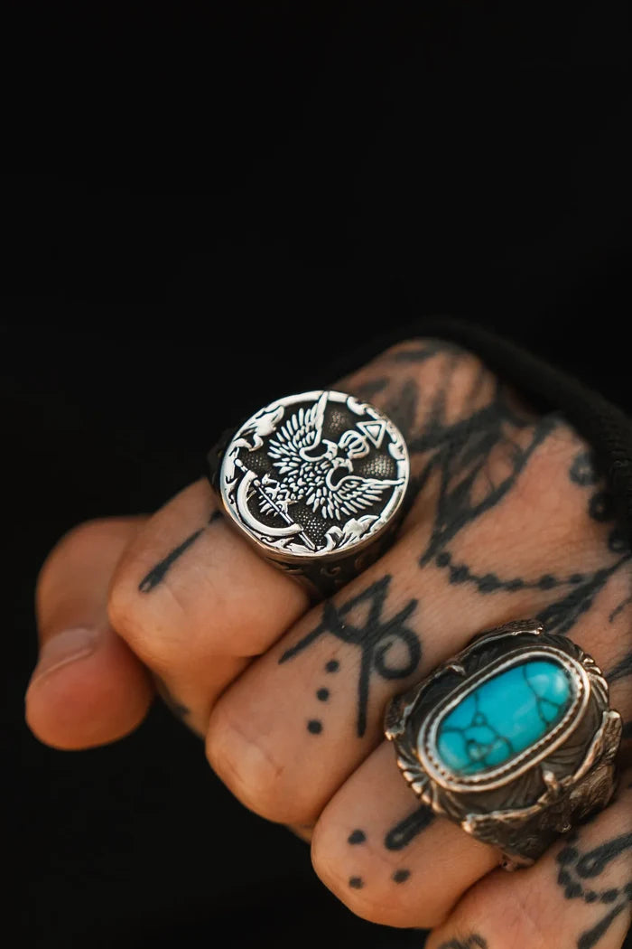 A tattooed hand wears two rings against a dark background: Relentless Betrayal’s handcrafted Revolution Ring with an eagle design, and another featuring a large turquoise stone.