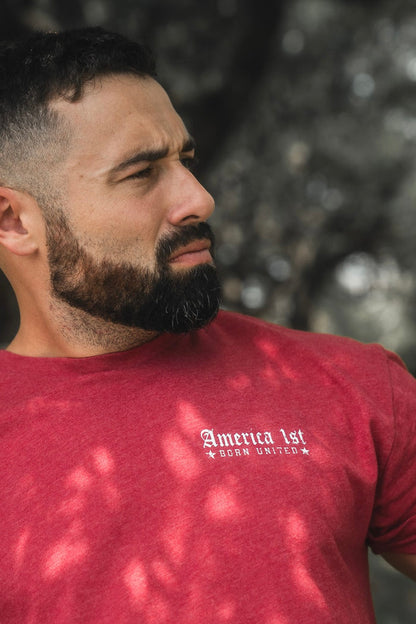 A bearded man with short hair, wearing a red "America First" t-shirt by Born United Private Label, looks to his left while standing outdoors with blurred trees in the background.