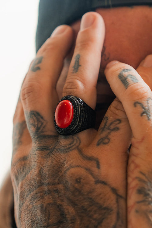 A tattooed hand adorned with Relentless Betrayal’s Burning Rose Blackout Ring, featuring a bold red stone set in black stainless steel, rests against a face as the detailed tattoos and striking ring catch the eye.