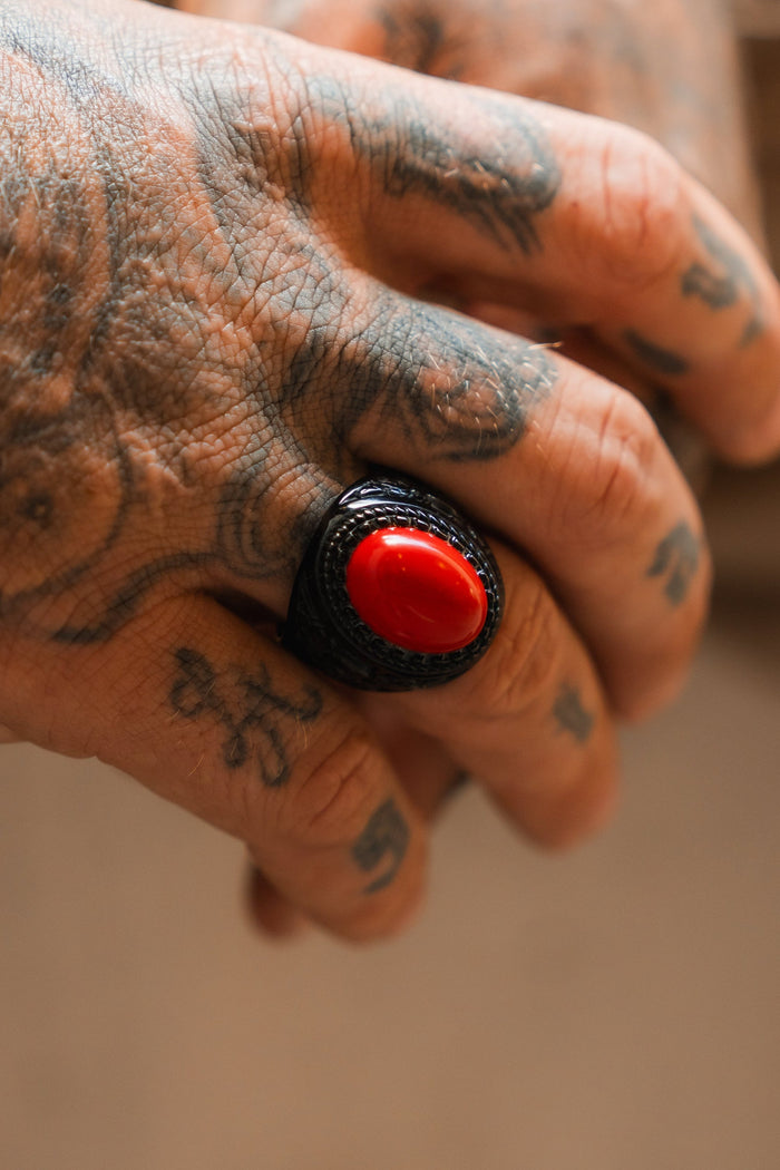 A tattooed hand showcases the Relentless Betrayal Burning Rose Blackout Ring, a handcrafted piece with a bold black band and striking red oval stone, intricate tattoos visible on the fingers and knuckles.