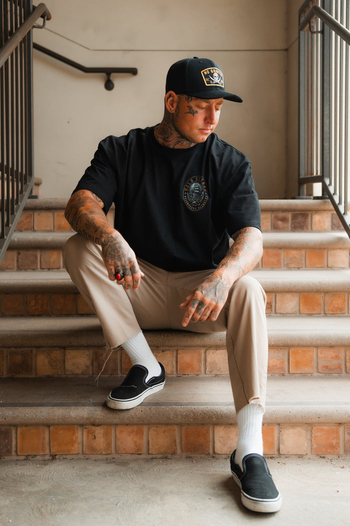 A tattooed man in a black cap and t-shirt, beige pants, and black slip-ons sits on stairs, wearing the Relentless Betrayal Burning Rose Blackout Ring, with one arm on his knee that shows off his tattoos.