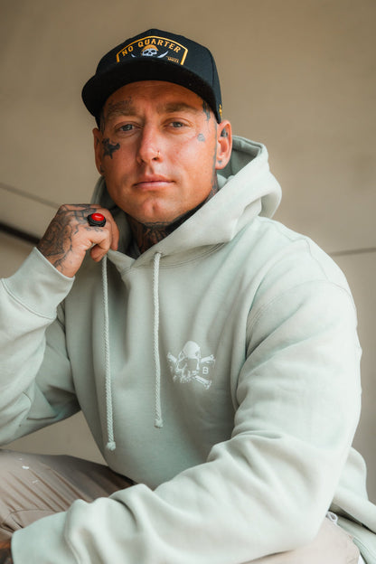 A tattooed person sits on indoor stairs, wearing a light gray hoodie, black NO QUARTER cap, and rests their chin on a hand featuring the Relentless Betrayal Burning Rose Blackout Ring.