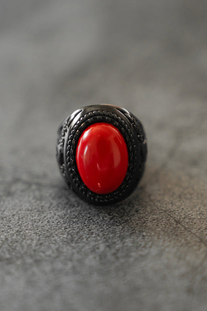 A close-up of the Relentless Betrayal Burning Rose Blackout Ring, a stainless steel piece with an ornate band and large oval red stone, is shown on a gray textured surface.