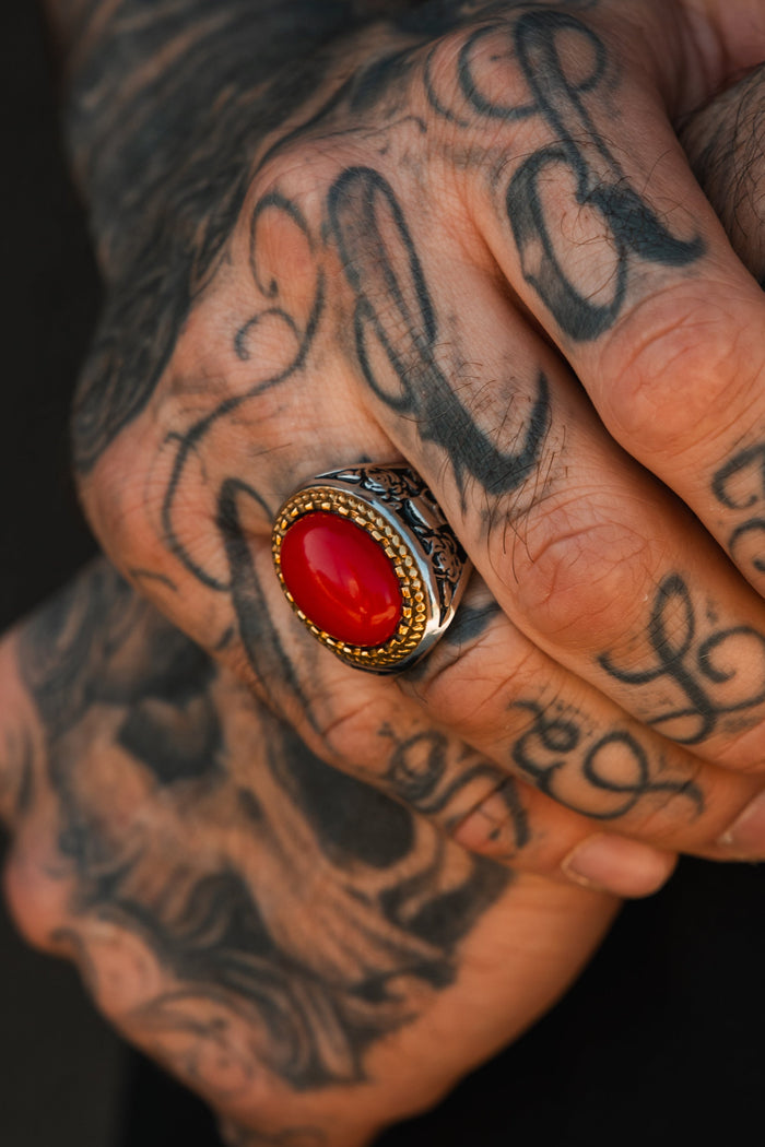 A close-up of tattooed hands, one adorned with the Relentless Betrayal Burning Rose Ring—a handcrafted piece featuring an ornate band and oval red stone. The bold black tattoos show swirling script and decorative patterns.