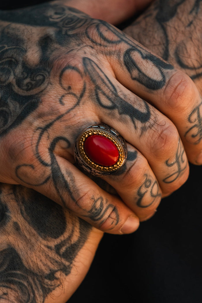 A close-up of tattooed hands with curled fingers, wearing the Burning Rose Ring by Relentless Betrayal—an artisanal statement piece featuring an oval red stone set in a detailed gold and silver band.