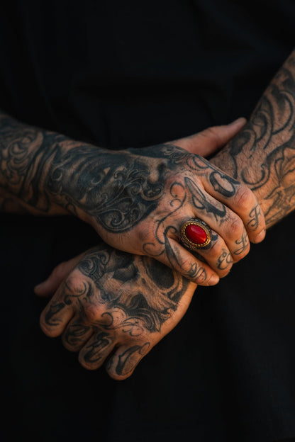 Close-up of two tattooed hands, one resting on the other, wearing the Relentless Betrayal Burning Rose Ring—a handcrafted ring featuring a striking red oval design. The person is dressed in black, with tattoos covering their fingers and hands.