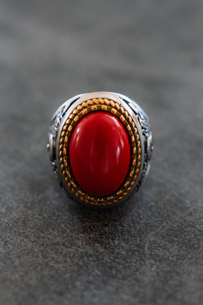 A close-up of the Burning Rose Ring by Relentless Betrayal, showcasing its ornate gold bezel and large smooth red oval stone at the center, displayed on a gray textured surface.