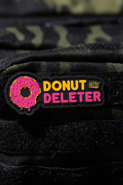 The Donut Deleter - Patch by Tango Charlie Apparel is a 3-inch 3D PVC patch with a pink frosted donut and "DONUT DELETER" text, Velcro-attached to the black front pocket of a camo backpack.