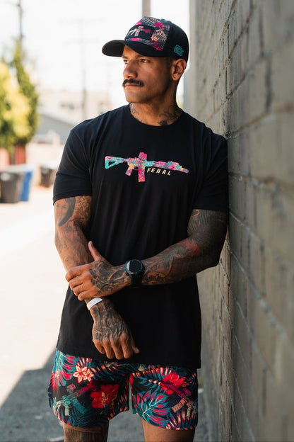 A tattooed man radiating radical authenticity wears the Feral - Tee by Tango Charlie Apparel—a black T-shirt with a colorful graphic—paired with a black cap and floral shorts as he leans against an outdoor brick wall on a sunny day.