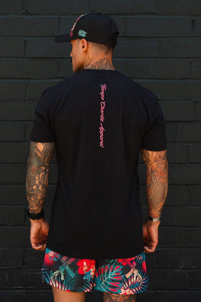 A man with tattooed arms stands before a black brick wall, wearing Tango Charlie Apparel's "Feral - Tee," a black T-shirt featuring pink text down the back, paired with a black cap and vibrant tropical shorts with red and green patterns.