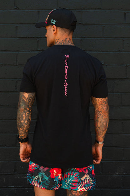 A man with tattooed arms stands before a black brick wall, wearing Tango Charlie Apparel's "Feral - Tee," a black T-shirt featuring pink text down the back, paired with a black cap and vibrant tropical shorts with red and green patterns.