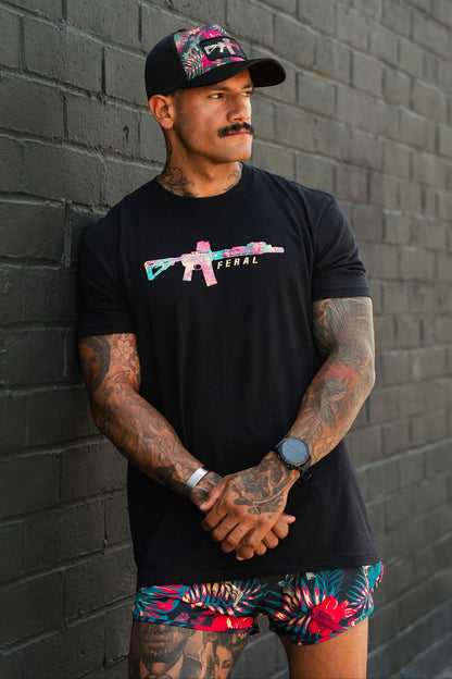 A tattooed man in a black cap stands against a brick wall, arms crossed. Wearing Tango Charlie Apparel's Feral - Tee with a vibrant gun graphic and floral shorts, he gazes to the side, embodying radical authenticity.