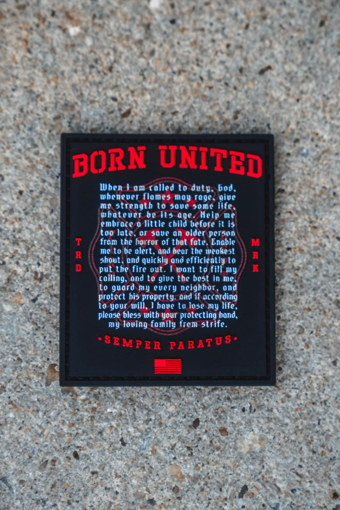 A Fireman's Prayer PVC Patch from Born United Private Label, featuring a black and red 3D design, rests on a gravel surface; it showcases the words "Born United," a prayer about duty and service, the motto "Semper Paratus," and an American flag graphic at the bottom.