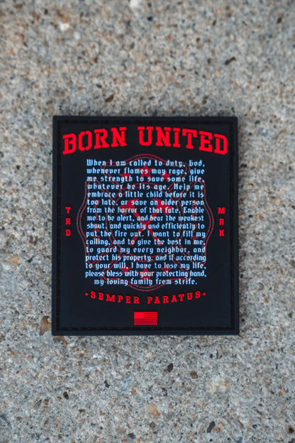 A Fireman's Prayer PVC Patch from Born United Private Label, featuring a black and red 3D design, rests on a gravel surface; it showcases the words "Born United," a prayer about duty and service, the motto "Semper Paratus," and an American flag graphic at the bottom.