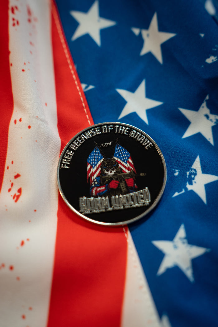 The Freedom Defender Challenge Coin by Born United Private Label, a round metal pin with skulls, American flags, and “Free Because Of The Brave,” rests on a red, white, and blue U.S. flag background.