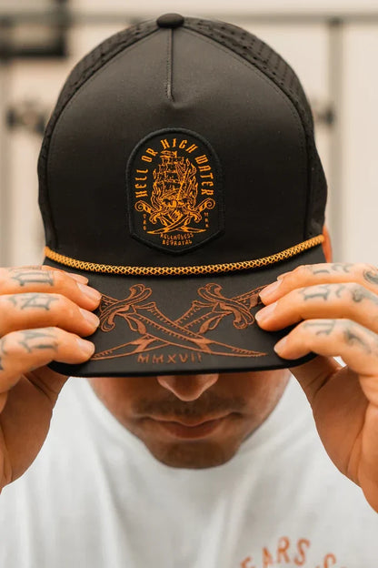 A person wears the Hell Or High Water Snapback by Relentless Betrayal, a black cap with an embroidered design and bold lettering. Tattooed fingers grip its visor, adorned with intricate patterns and Roman numerals MMXVII, reflecting resilience themes.