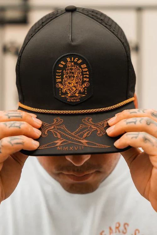 A person wears the Hell Or High Water Snapback by Relentless Betrayal, a black cap with an embroidered design and bold lettering. Tattooed fingers grip its visor, adorned with intricate patterns and Roman numerals MMXVII, reflecting resilience themes.