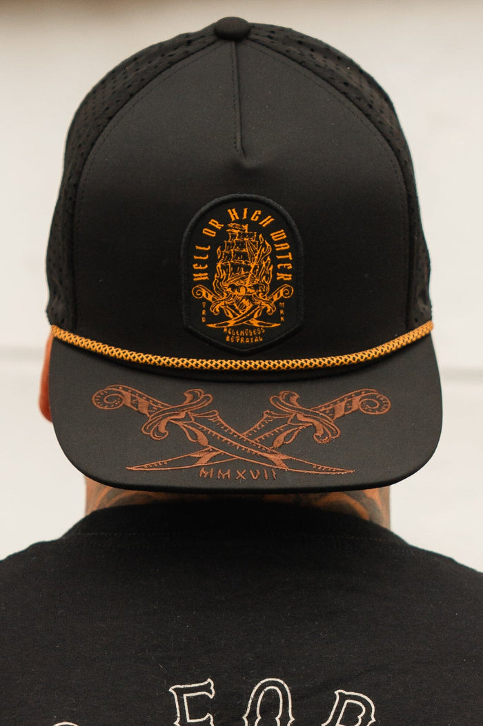 A person wears the Hell Or High Water Snapback by Relentless Betrayal, a black cap with gold embroidery displaying a ship and Hell or High Water on the front. The cap includes an anchor design and MMXVII Roman numerals on the brim, highlighting resilience.