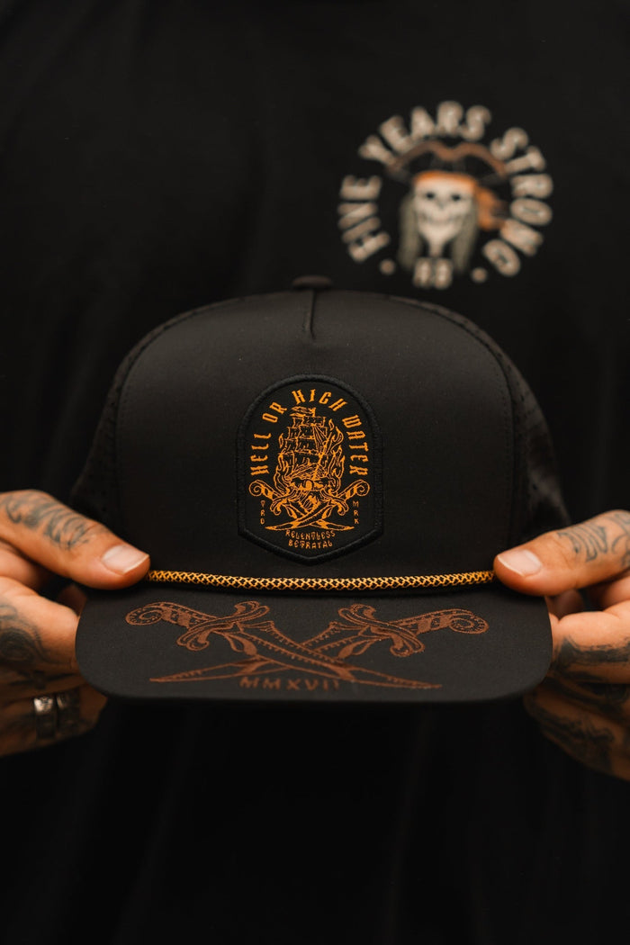 A person with tattooed hands holds a Hell Or High Water water-resistant snapback from Relentless Betrayal, showcasing a gold embroidered emblem and text. In a black shirt adorned with a pirate skull and Five Years Strong, they exude resilience.