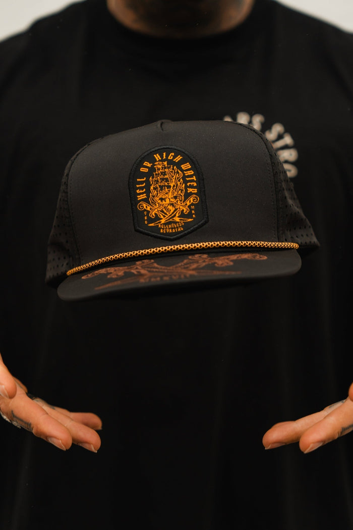 A person holds the Relentless Betrayal black Hell Or High Water snapback adorned with an orange emblem and gold chain. The blurred background highlights this bold pieces resilience.