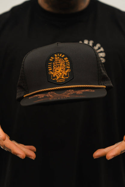 A person holds the Relentless Betrayal black Hell Or High Water snapback adorned with an orange emblem and gold chain. The blurred background highlights this bold pieces resilience.