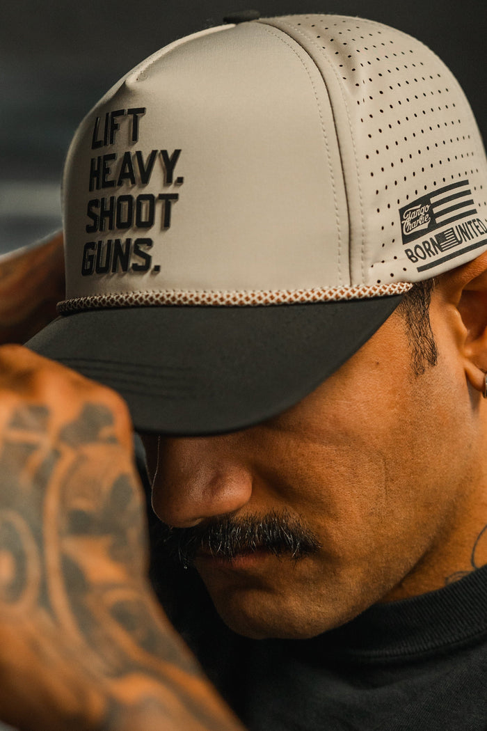 A tattooed man with a mustache adjusts the brim of his gray and black LHSG Hydro Snapback Hat by Tango Charlie Apparel, featuring “LIFT HEAVY, SHOOT GUNS” from the Born United collection.