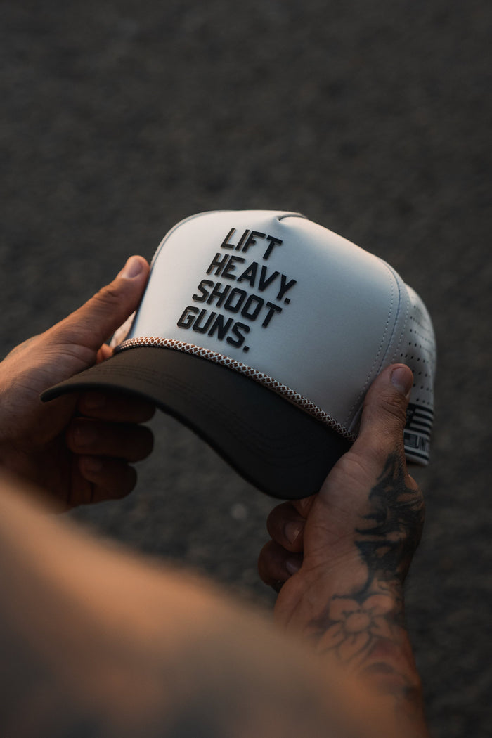 A tattooed hand holds the LHSG Hydro - Snapback Hat by Tango Charlie Apparel, a hydro-resistant white and black snapback with "LIFT HEAVY. SHOOT GUNS." text, shown against a dark textured background.
