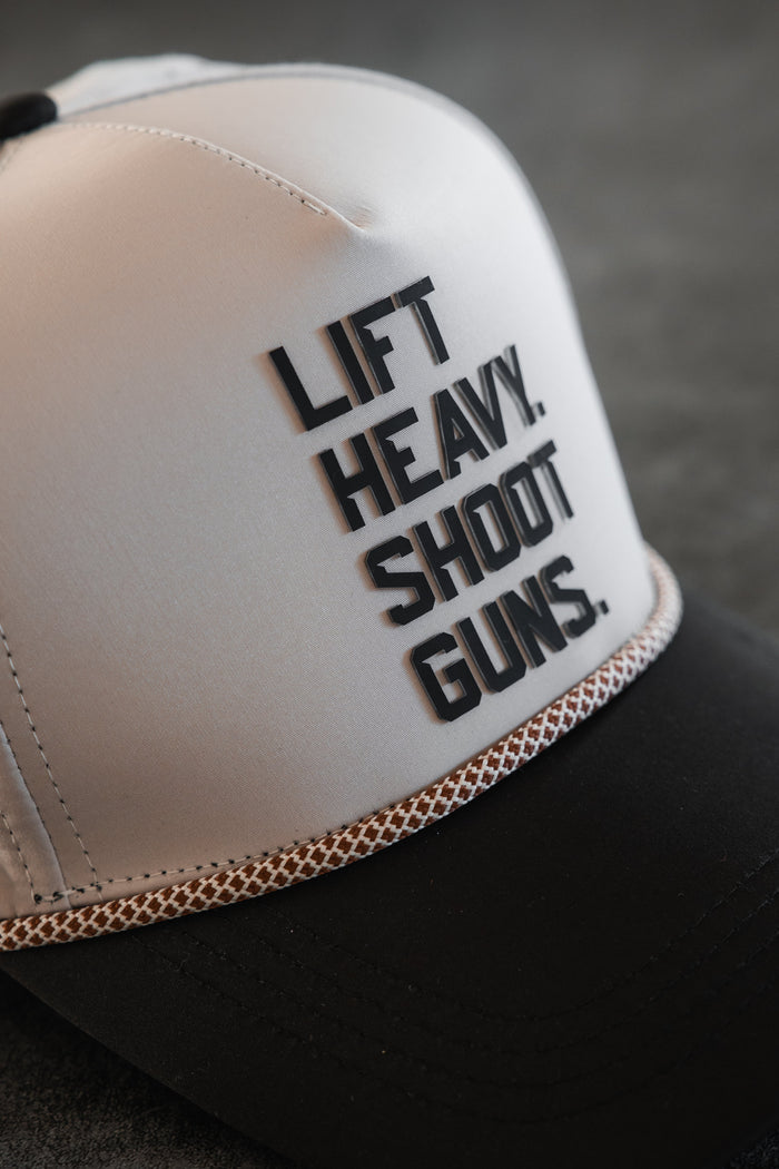 Close-up of the LHSG Hydro Snapback Hat by Tango Charlie Apparel, featuring a white front, black bill, braided trim, and raised black “LIFT HEAVY. SHOOT GUNS.” text—a stylish two-tone cap from the Born United collaboration.