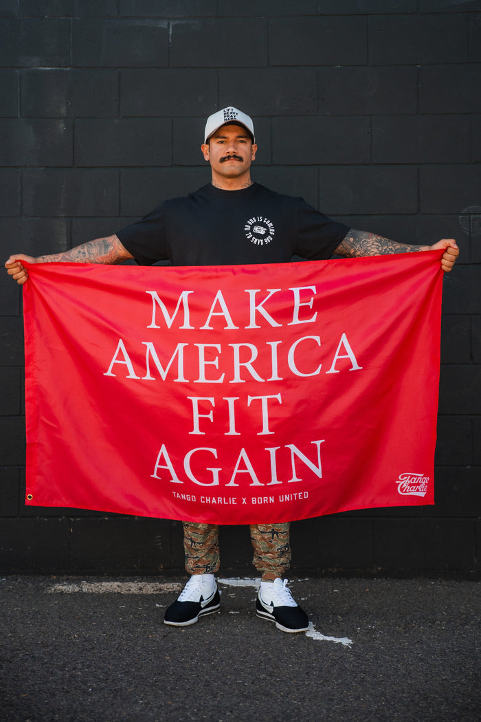 A tattooed man in a black t-shirt, white cap, and sneakers holds the Tango Charlie Apparel "Make America Fit Again" Flag against a black wall.
