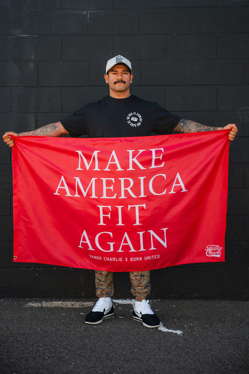 A tattooed man in a black t-shirt, white cap, and sneakers holds the Tango Charlie Apparel "Make America Fit Again" Flag against a black wall.
