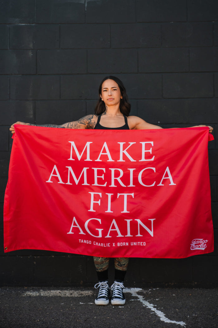 A tattooed woman stands before a black brick wall, holding the bold polyester "Make America Fit Again - Flag" by Tango Charlie Apparel.
