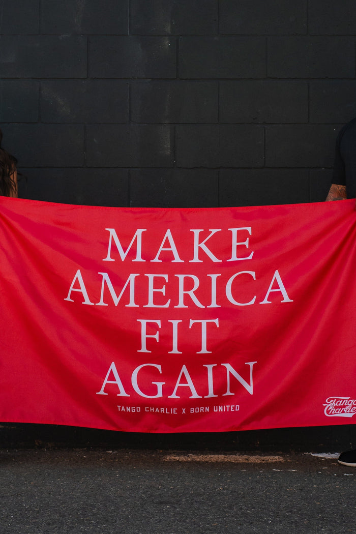 Two people in camo pants and black tops hold the Tango Charlie Apparel "Make America Fit Again" red polyester flag in front of a dark wall. The banner features bold white lettering.