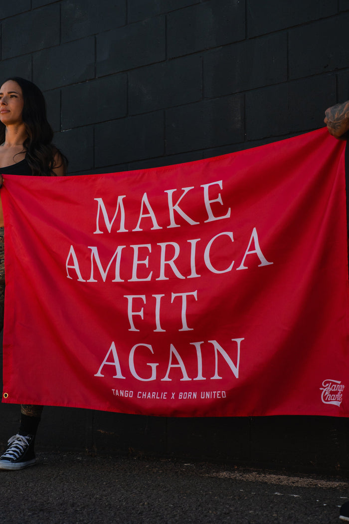 Two people partially visible hold the Make America Fit Again - Flag by Tango Charlie Apparel, a red polyester banner with white text reading “MAKE AMERICA FIT AGAIN. TANGO CHARLIE X BORN UNITED,” against a dark brick wall.