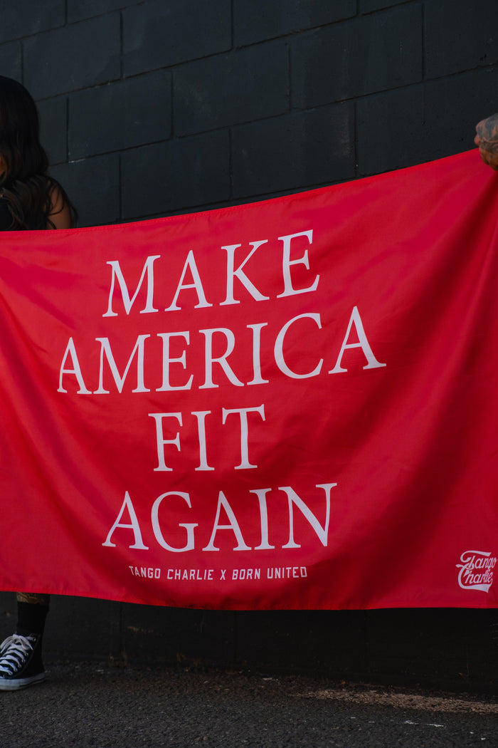 A person holds the Tango Charlie Apparel Make America Fit Again - Flag, featuring bold white text on red polyester, against a dark brick wall. Only part of their arm and shoe are visible as they grip this striking gym banner.