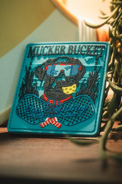 Add playful flair to your gear with the Pickleball Retriever PVC Patch by Mucker Bucket—a vibrant 4x4 Velcro patch featuring a dog sporting sunglasses, a red collar, and a yellow pickleball in its mouth, along with "Mucker Bucket" and "Pickleball Club" text. Perfect for customizing your accessories!.