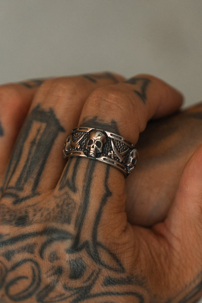 A close-up of a tattooed hand wearing the Relentless Betrayal Piracy Ring, featuring bold skull and wing details. The blurred background highlights the intricate tattoos and the standout ring design.