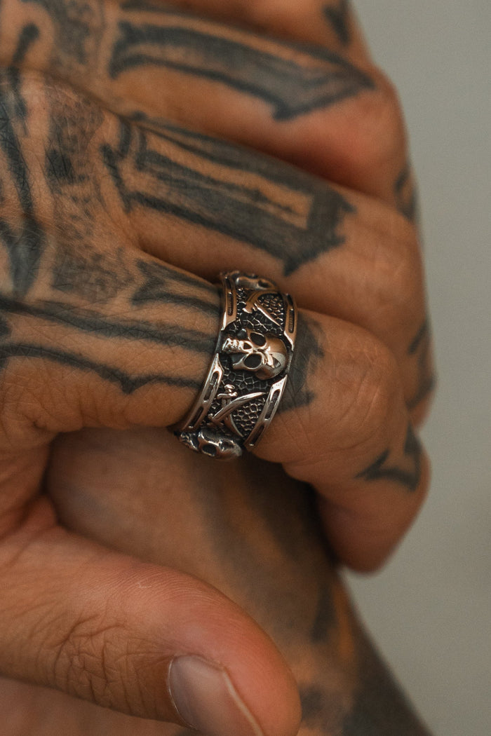 A hand with geometric black tattoos wears the Piracy Ring by Relentless Betrayal, a detailed stainless steel skull piece. The softly curled fingers are set against a neutral background.