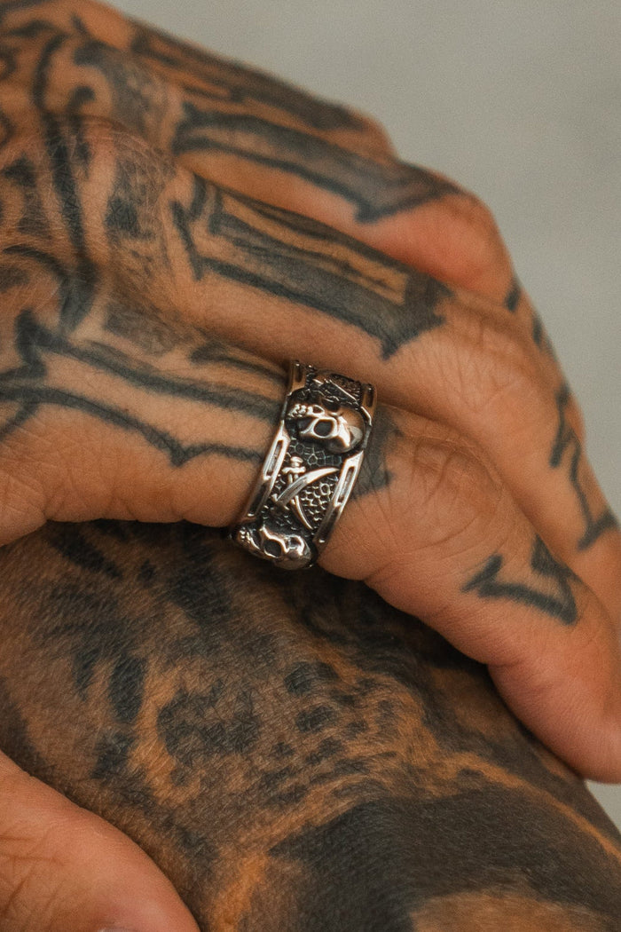 A close-up of a tattooed hand wearing the Piracy Ring by Relentless Betrayal—a thick stainless steel skull ring with intricate detailing—resting gently on another tattooed surface.