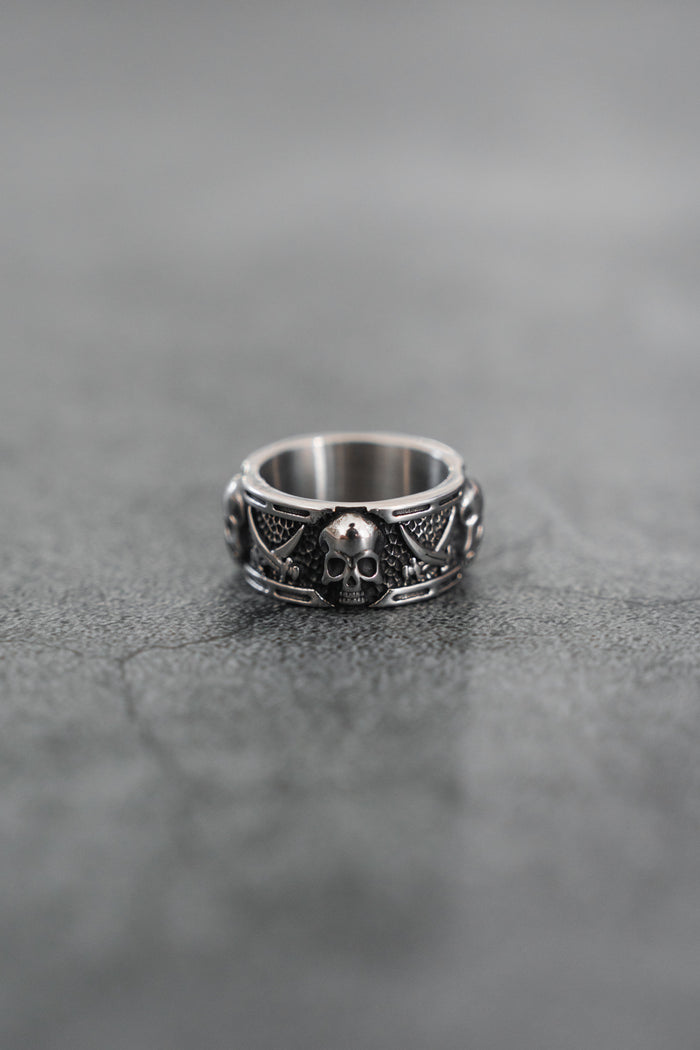 The Piracy Ring by Relentless Betrayal features a bold stainless steel skull on the front, accented by intricate patterns and smaller skulls along the band, all set against a textured gray surface for a striking look.