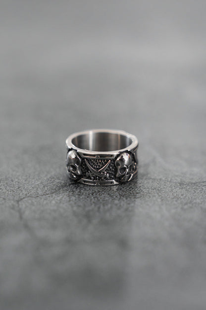 The Piracy Ring by Relentless Betrayal is a stainless steel piece with intricate details, showcasing two skulls and a triangular pattern, displayed on a textured gray surface.
