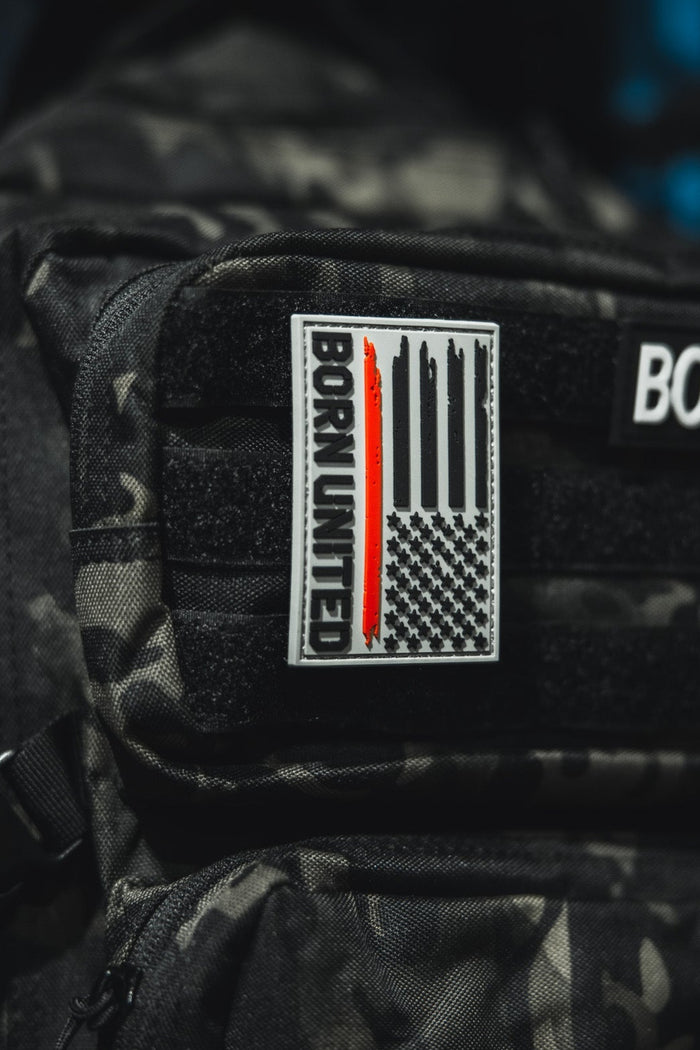 A close-up of a black camo backpack displays the Red Line Flag PVC Patch by Born United Private Label, featuring a grayscale American flag with a single red stripe and bold BORN UNITED text.