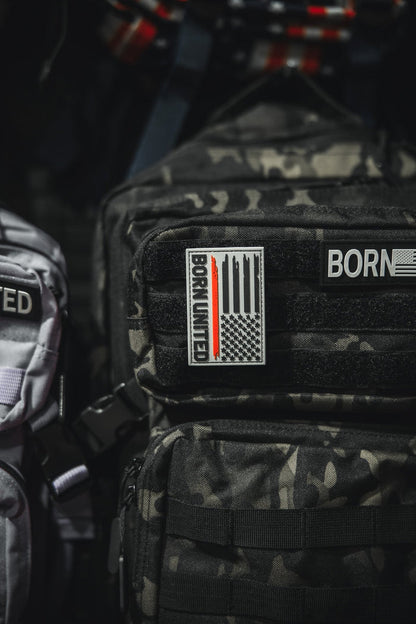 A black camouflage backpack displays a Red Line Flag PVC Patch by Born United Private Label with a stylized U.S. flag and BORN UNITED text. Above it is another partial BORN patch. A light gray bag appears partially on the left.