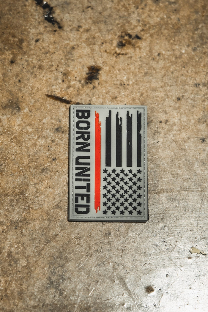 The Red Line Flag PVC Patch by Born United Private Label is a rectangular patch with a stylized American flag, featuring one red stripe and “BORN UNITED” vertically on the left, displayed on a worn, brownish metal surface.