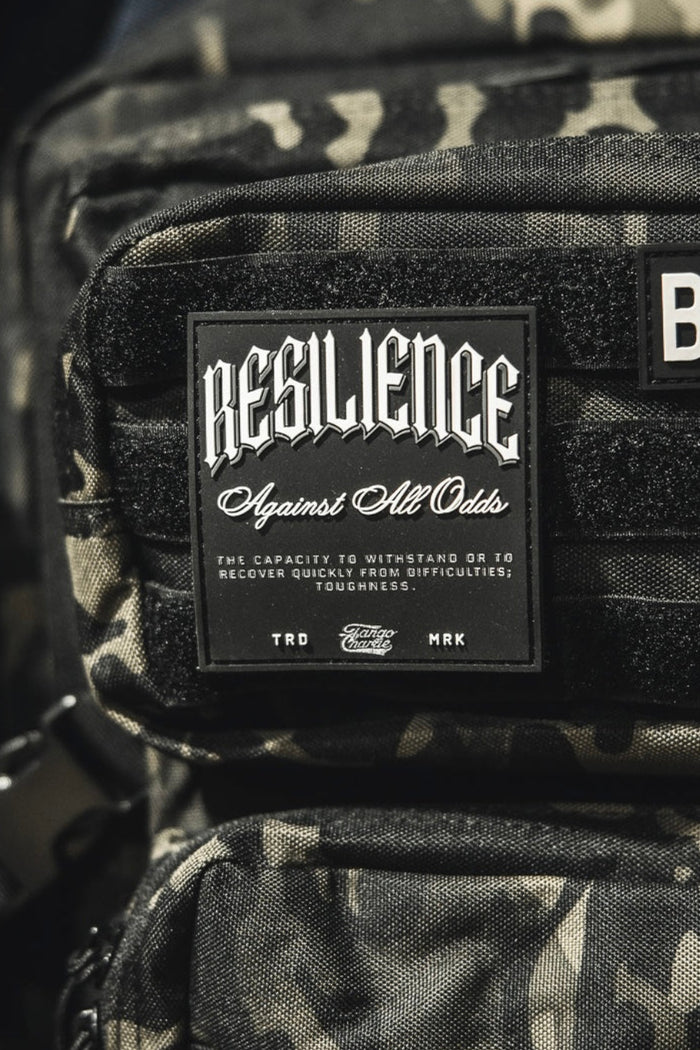 A close-up shows a camouflage backpack with the Tango Charlie Apparel "Resilience - Patch" and a 3D PVC American flag patch reading BORN. Part of a pink bag appears on the left.