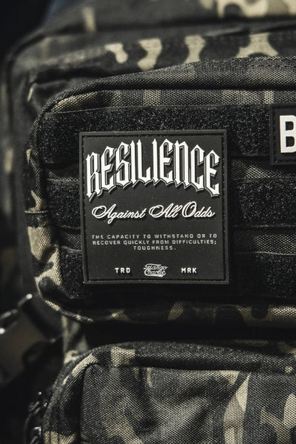 A close-up shows a camouflage backpack with the Tango Charlie Apparel "Resilience - Patch" and a 3D PVC American flag patch reading BORN. Part of a pink bag appears on the left.