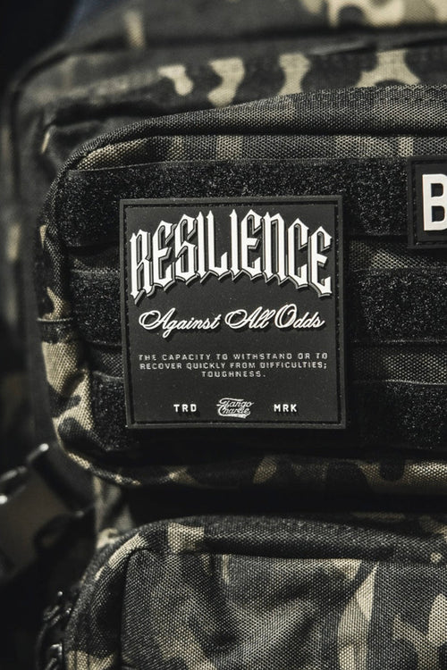 A close-up shows a camouflage backpack with the Tango Charlie Apparel "Resilience - Patch" and a 3D PVC American flag patch reading BORN. Part of a pink bag appears on the left.