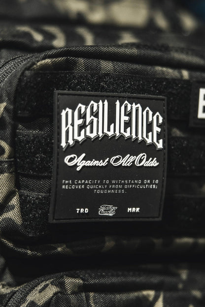 A close-up of the Tango Charlie Apparel "Resilience - Patch," a 3D PVC patch on a camo bag, featuring the phrase "RESILIENCE Against All Odds" to symbolize toughness and overcoming challenges.