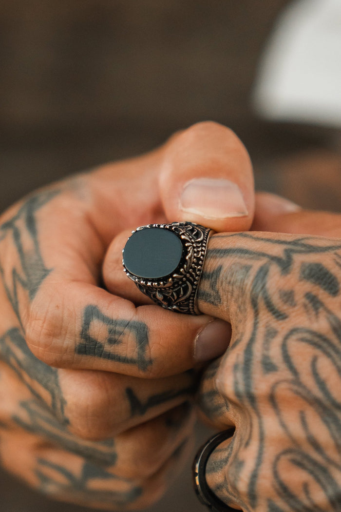 A close-up of tattooed hands adjusting the Relentless Betrayal Royal Darkness Ring, showcasing its striking black finish, intricate stainless steel metalwork, and bold, elegant style.
