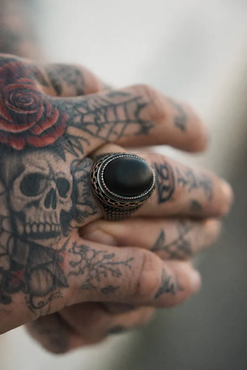 A tattooed hand adorned with a skull and rose design wears The Dark Current Ring by Relentless Betrayal—a large, handcrafted men's ring showcasing an oval black stone on the ring finger.